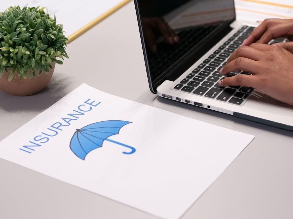 A person typing on a laptop with an insurance print-out sitting on a desk.